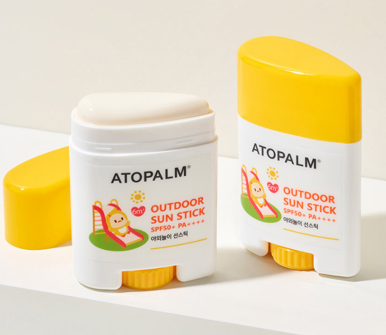 Two Atopalm Outdoor Sun Stick SPF50+ PA++++ stick containers on a white surface with a light beige background. One container is open, showing the cream-colored stick, and the other is closed.