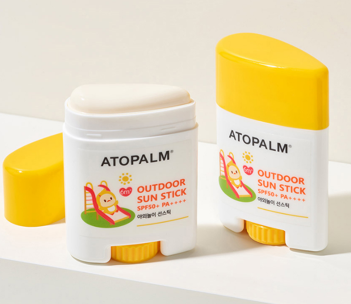 Two Atopalm Outdoor Sun Stick SPF50+ PA++++ stick containers on a white surface with a light beige background. One container is open, showing the cream-colored stick, and the other is closed.